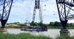 Visiteurs traversant le pont transbordeur de Rochefort, avec des oiseaux volant autour et des paysages verdoyants en arrière-plan.